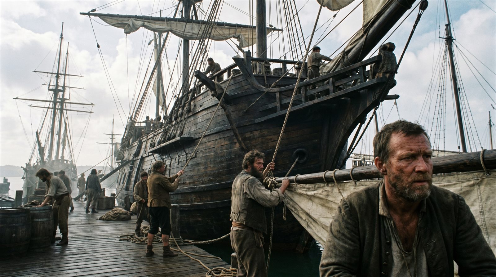 Dockworkers hauling rope beside a tall wooden sailing ship moored at a Portuguese quay under an overcast sky.